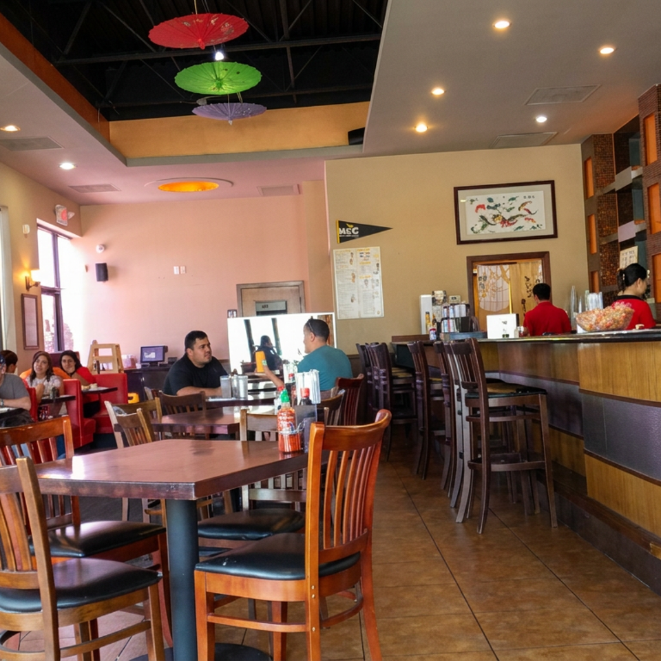 Bright Asian restaurant interior with diners, wooden tables, red booths, colorful ceiling decorations, and a counter adorned with lucky cat figurines.